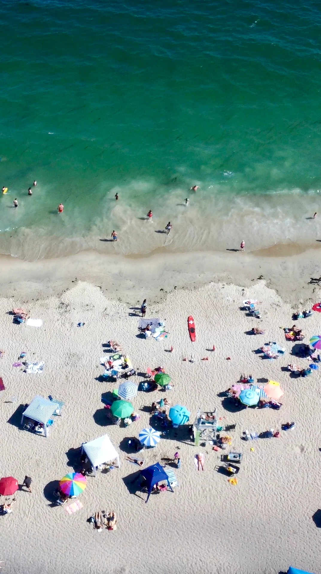 An aerial view of turquoise surf meeting a sandy beach dotted with colorful umbrellas and beachgoers