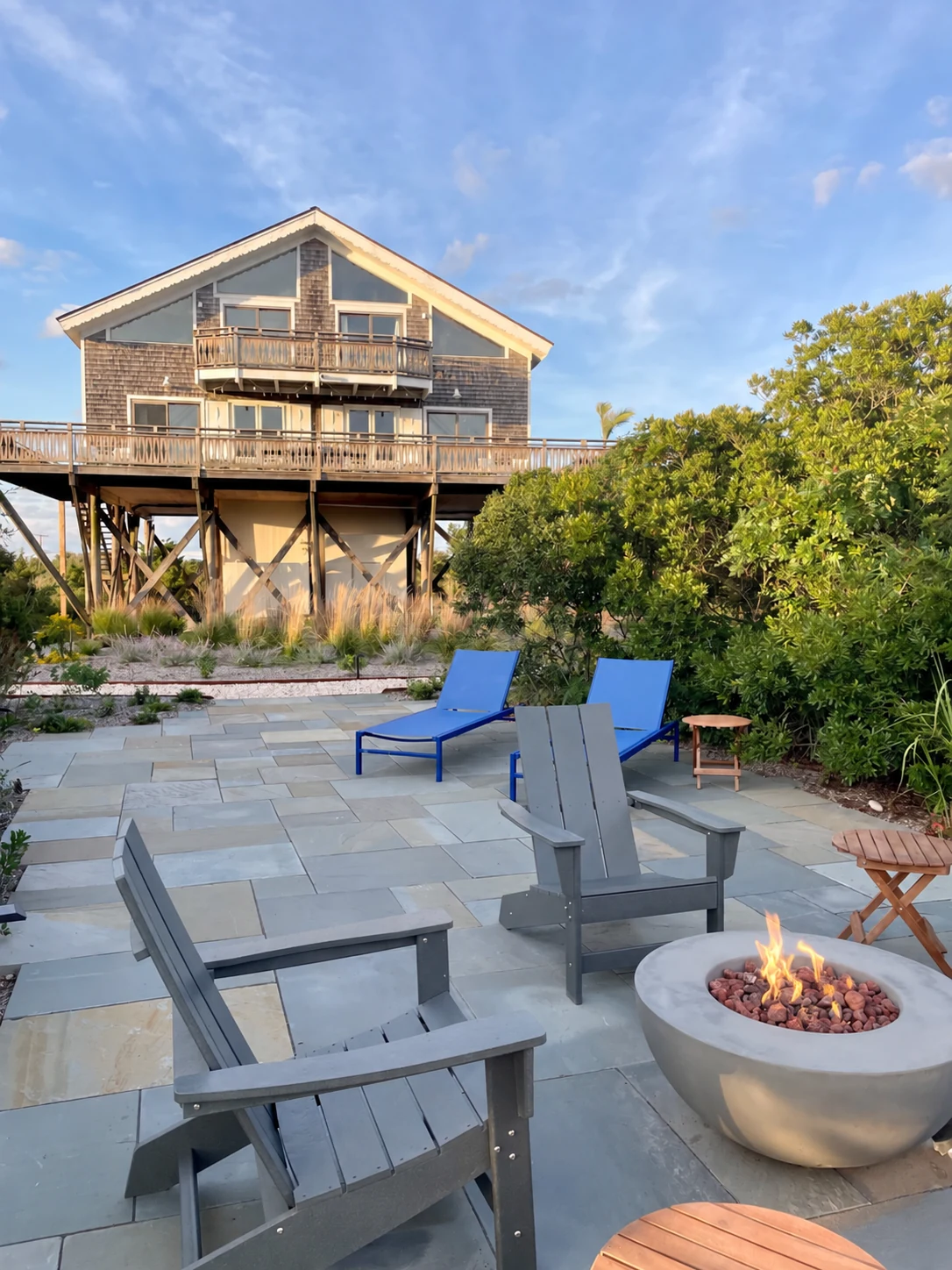 A lit fire pit with gray Adirondack chairs and blue loungers on the sunlit Surf House patio